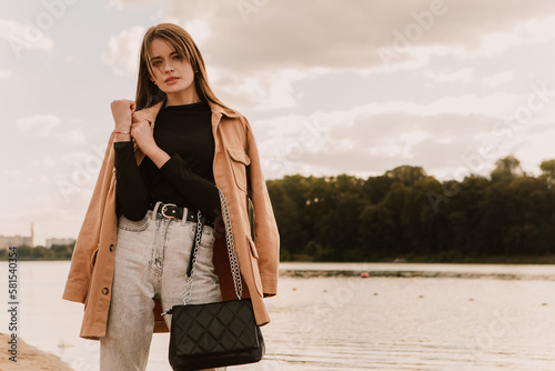 Caucasian young pretty stylish woman standing near river lake water street on sand pier dressed brown trench coat hold handbag smiling poses outside city, spring autumn season. Cute brunette hair lady