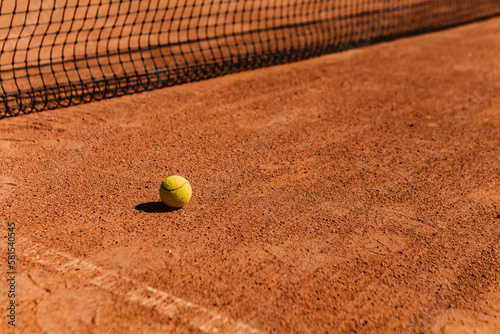 Light orange empty outdoor clay court surface dry grungy ground baseline detail for playing tennis with net in sunny day with yellow green ball, gravel texture background, copy space for text    © Volodymyr