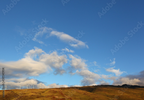 Wind Turbines in a row along a hillside against a wide blue sky with fluffy clouds