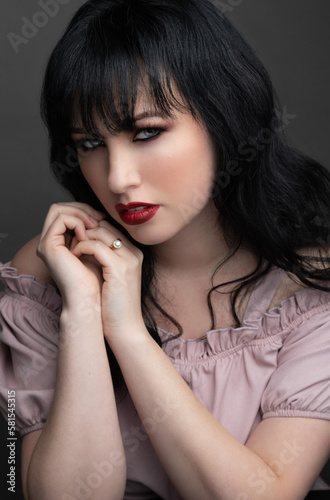 Close up portrait of a caucasian woman in her 30s. She is wearing a pink dress and red lipstick. She has black hair and blue eyes. This is a studio portrait with a grey background.