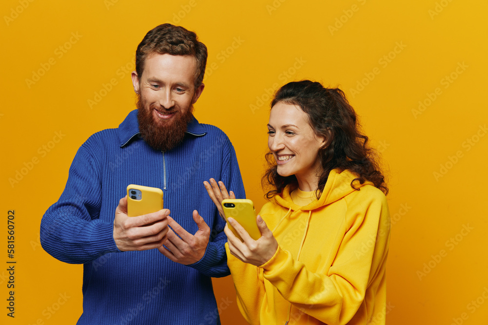 Man and woman couple smiling merrily with phone in hand social media viewing photos and videos, on yellow background, symbols signs and hand gestures, family freelancers. Man and woman couple smiling merrily with phone in hand social media viewing photos and videos, on yellow background, symbols signs and hand gestures, family freelancers.