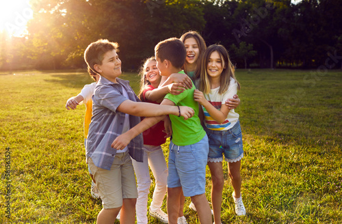 Cheerful children girls and boys hugging each other on sunny lawn during summer walk with school friends or classmates rejoicing at start of holidays, dressed in casual kids clothes standing in park
