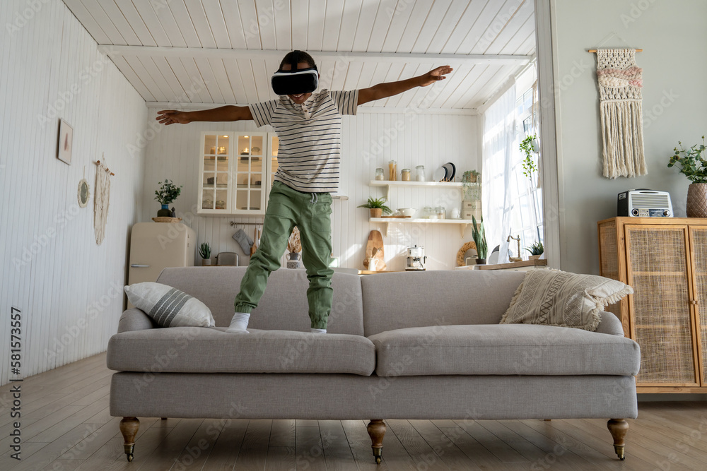 Carefree African boy child using VR helmet for fun and entertainment, happy kid standing on sofa playing immersive 3D virtual flight game, flying in metaverse world. Children in immersive environment - obrazy, fototapety, plakaty 