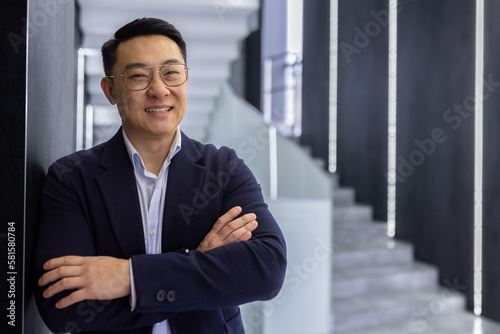 Portrait of successful asian businessman inside office building, man with crossed arms smiling and looking at camera, boss in business suit and glasses standing in hall corridor of office center.