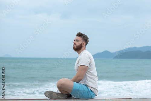 Young calm sad serious man is sitting on embankment near sea, ocean on beach, thinking, meditate on natural background 