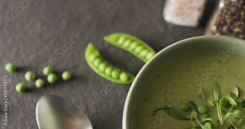 Close up of white bowl of pea soup with spoon, peas and seasoning on dark background