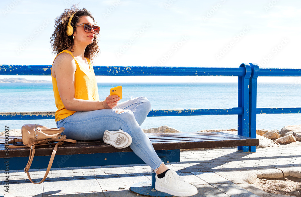 Female meloman resting on bench on seashore in sunshine
