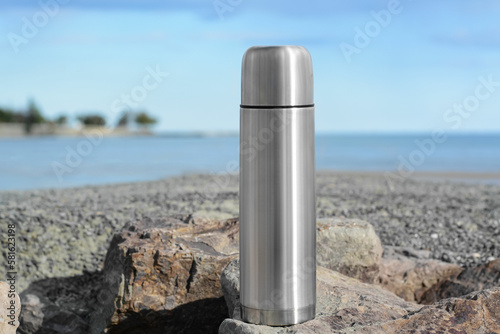 Metallic thermos with hot drink on stone near sea