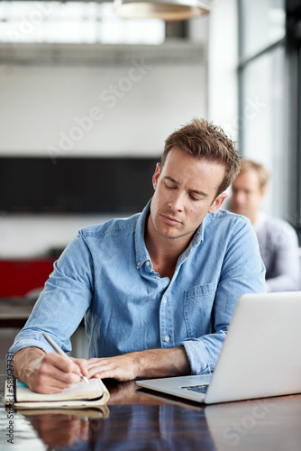 Staying connected to his creativity. a man writing notes while working on a laptop in an office.