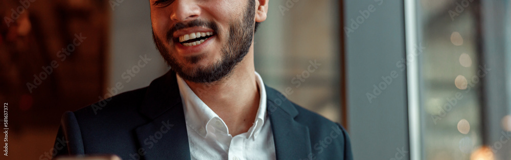 Obraz premium Smiling Indian businessman wearing suit is using phone during lunch time in cafe