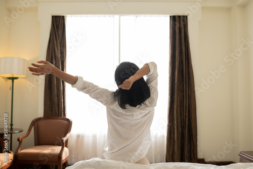 Asian woman wake up in the morning, sitting on white bed and stretching, feeling happy and fresh. happy woman lying on soft pillows awaken in cozy bedroom, relaxation concept.