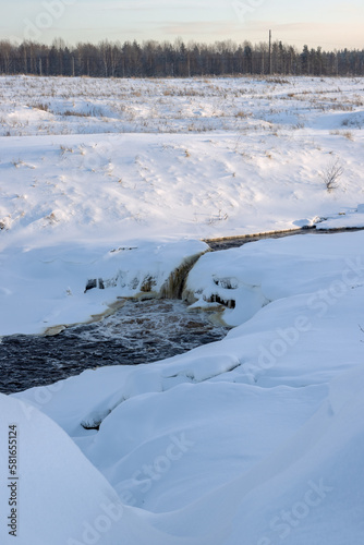 Fototapeta Naklejka Na Ścianę i Meble -  Top view of an icy freezing waterfall. Winter river landscape. Stormy streams of water among ice and snow. Cold winter weather. Tosnensky waterfall, Tosna river, Ulyanovka, Leningrad Oblast, Russia.