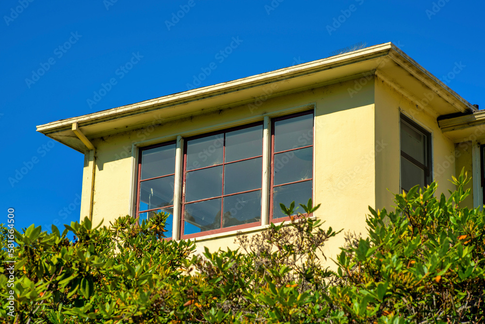 Visible room or apartment with flat roof and yellow cement exterior with front or back yard trees and blue skyline