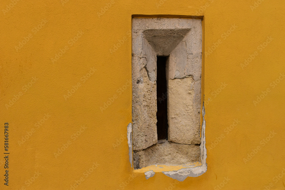 yellow stucco exterior wall elevation detail with rough stone window. narrow arrow slit with bevel. restored old stone wall. historic building closeup. restoration and renovation method and concept. yellow stucco exterior wall elevation detail with rough stone window. narrow arrow slit with bevel. restored old stone wall. historic building closeup. restoration and renovation method and concept.
