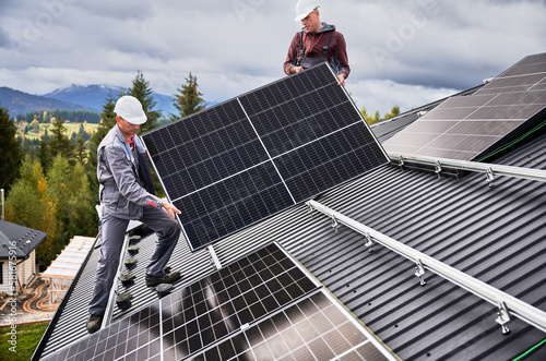 Engineers building solar panel system on roof of house. Men workers in helmets carrying photovoltaic solar module outdoors. Concept of alternative and renewable energy. © anatoliy_gleb