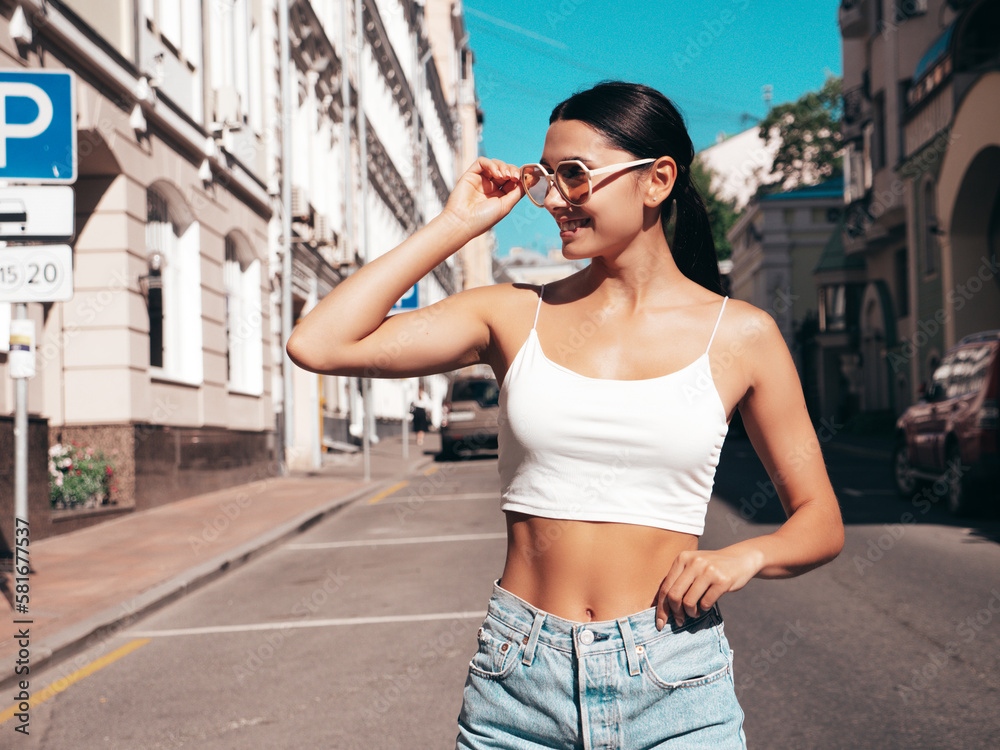 Young beautiful smiling hipster woman in trendy summer white top and shorts clothes. Sexy carefree model posing on the street background at sunny day. Positive brunette female. In sunglasses Young beautiful smiling hipster woman in trendy summer white top and shorts clothes. Sexy carefree model posing on the street background at sunny day. Positive brunette female. In sunglasses
