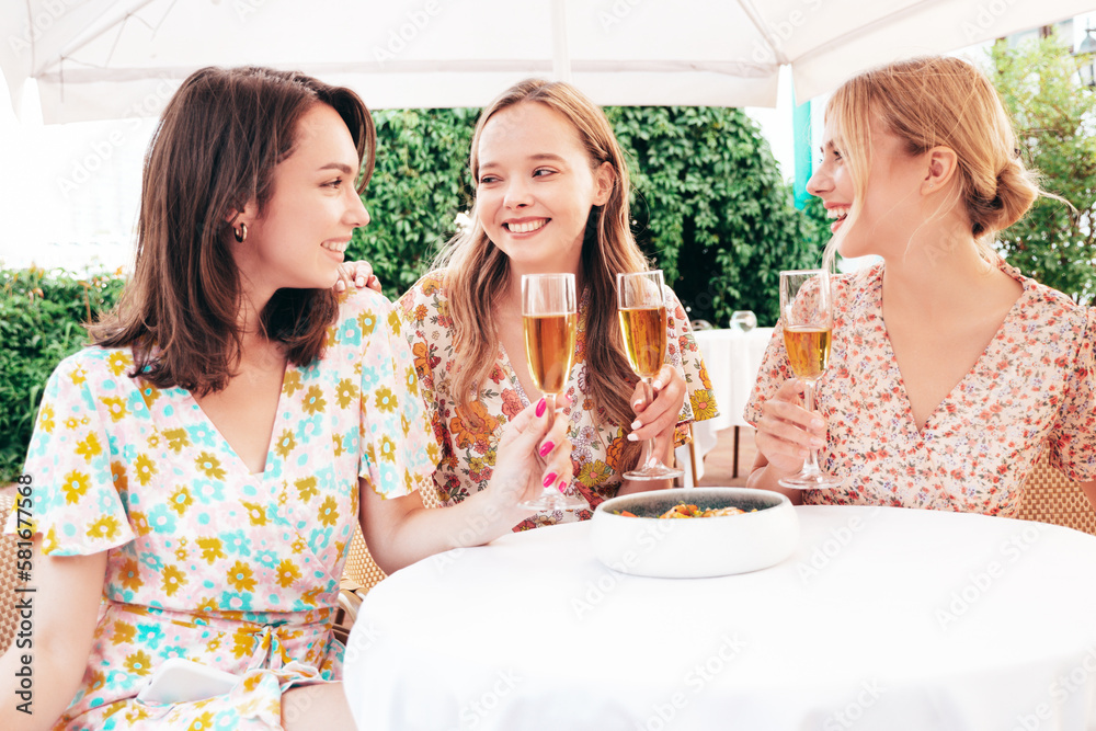 Three young beautiful smiling hipster female celebrating. Carefree women posing at summer terrace veranda cafe in the street.Positive models drinking champagne. Enjoying their vacation. Talking