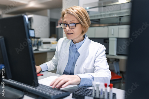 Female Scientists Working on their Computers In Big Modern Laboratory. Various Shelves with Beakers, Chemicals and Different Technical Equipment is Visible. photo