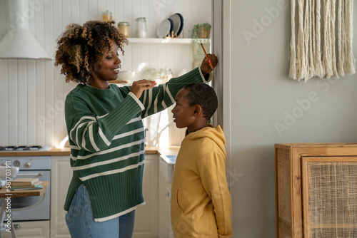 African American stay-at-home mom measuring height of kid son at home, school-aged boy standing against wall while mother taking measurement with ruler and pencil. Child growth and development concept photo