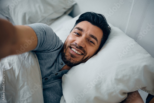 Happy man taking selfie while relaxing in bedroom and looking at camera.
