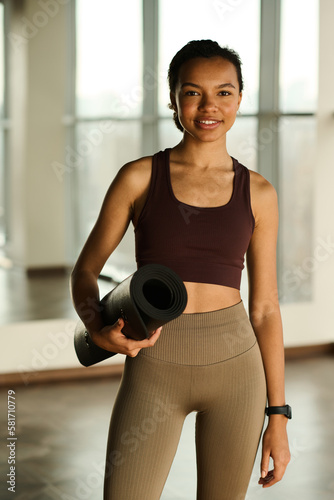 Portrait of young healthy girl with exercise mat smiling at camera while standing in health club