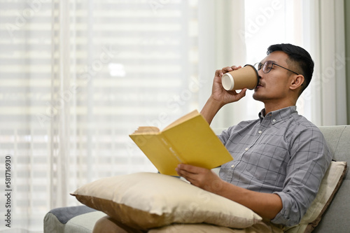Bilde på lerret Side view of a smart Asian man sipping coffee and reading book while relaxing in living room