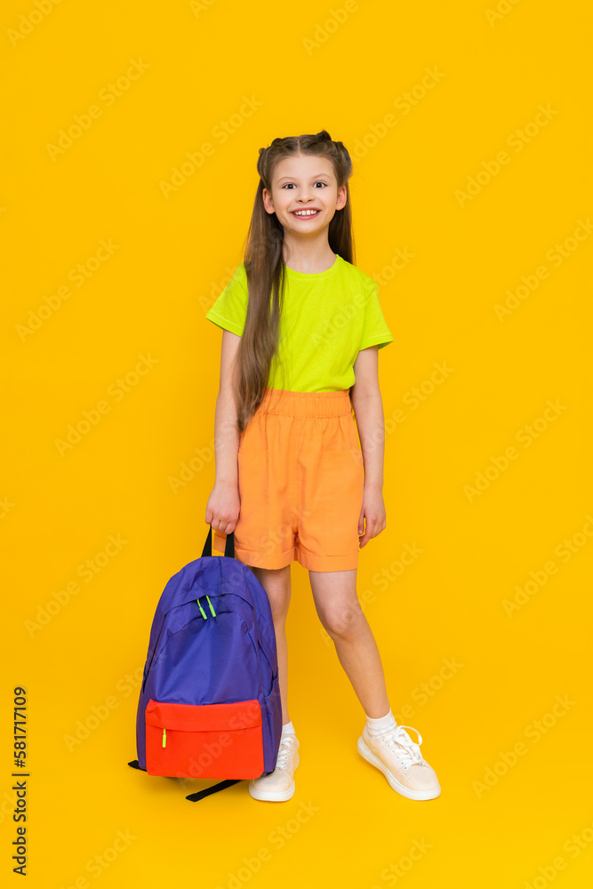 A little girl with a school backpack. A child in summer shorts is going to educational courses. A happy child with a satchel. A full-length schoolgirl on a yellow isolated background. A little girl with a school backpack. A child in summer shorts is going to educational courses. A happy child with a satchel. A full-length schoolgirl on a yellow isolated background.