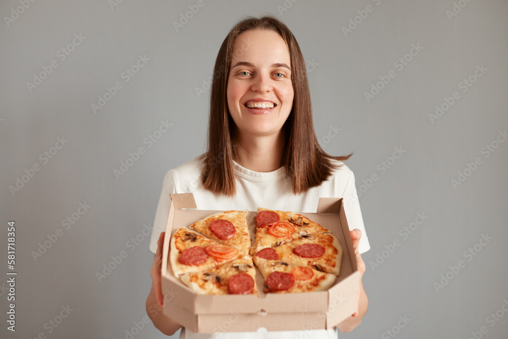 Image of pretty dark haired woman wearing white casual t-shirt smiling and holding pizza box isolated over gray background, ordering fast food delivery for dinner. Image of pretty dark haired woman wearing white casual t-shirt smiling and holding pizza box isolated over gray background, ordering fast food delivery for dinner.