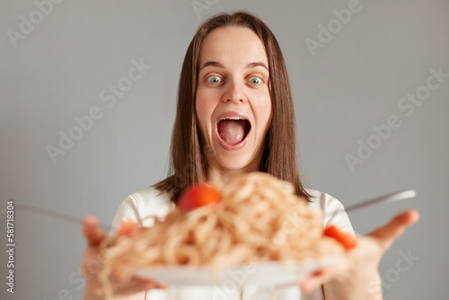 Indoor shot of amazed excited woman with brown hair taking big plate with tasty pasta isolated over gray background  being extremely hungry  keeps mouth widely opened.