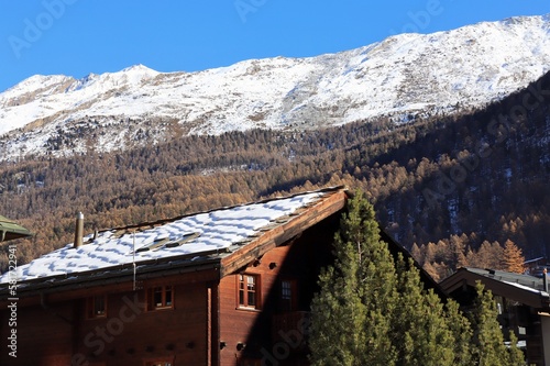 Wooden house with snow covered roof in winter. There is a Swiss Alps with snow capped peaks in the background.