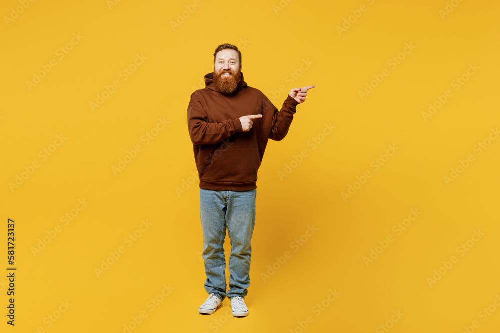 Full body young redhead caucasian man wearing brown hoody casual clothes pointing indicate on workspace area copy space mock up isolated on plain yellow background studio portrait. Lifestyle concept. Full body young redhead caucasian man wearing brown hoody casual clothes pointing indicate on workspace area copy space mock up isolated on plain yellow background studio portrait. Lifestyle concept.
