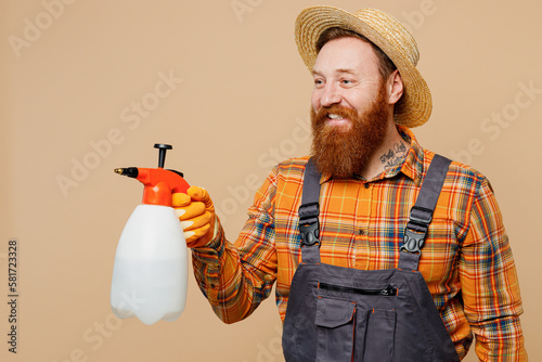 Happy fun young bearded man wear straw hat overalls work in garden hold in hand use chemical spray in bottle isolated on plain pastel light beige color background studio portrait Plant caring concept