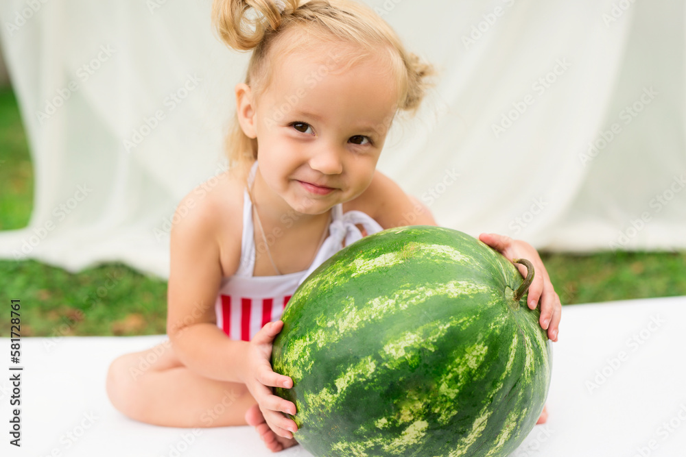 Fototapeta premium Little girl with a big watermelon on a white mattress in the garden