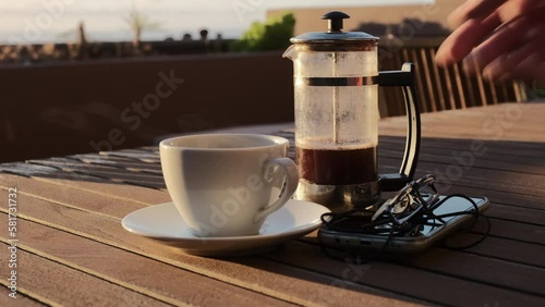 Café caliente humeante de cafetera de émbolo sobre una mesa de madera con luz de amanecer en una terraza con vistas al mar y jardinera de fondo, con mano de hombre que sirve café. photo