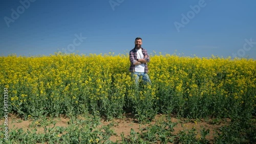 Happy and successful farmer is standing by his rapeseed field. Rapeseed plantation in bloom. Slow motion video.
