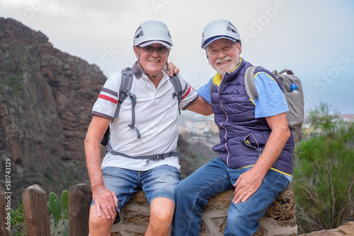 Senior couple of men hikers with helmets and backpacks resting on the top of mountain - Smiling climbing tourists enjoying holidays and healthy lifestyle in trekking day