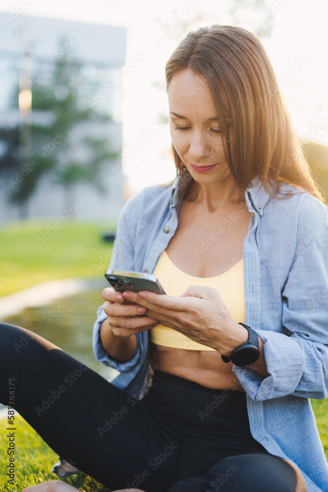 Woman in sportswear, after jogging in the park, is resting, using phone or chatting on a mobile phone. Holding smartphone. Sitting woman outdoors. Woman in sportswear, after jogging in the park, is resting, using phone or chatting on a mobile phone. Holding smartphone. Sitting woman outdoors.