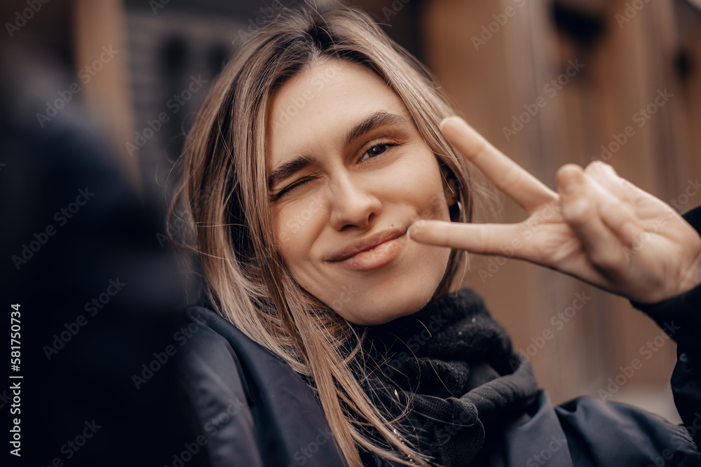 Stylish girl with blonde hair making selfie outside. Cool woman in fashion bomber showing peace sign and smiling outdoors.