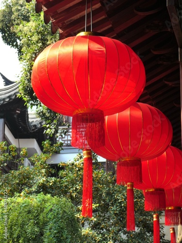 Photography Red Chinese Lanterns hanging from a wooden roof with Chinese buildings and greenery behind