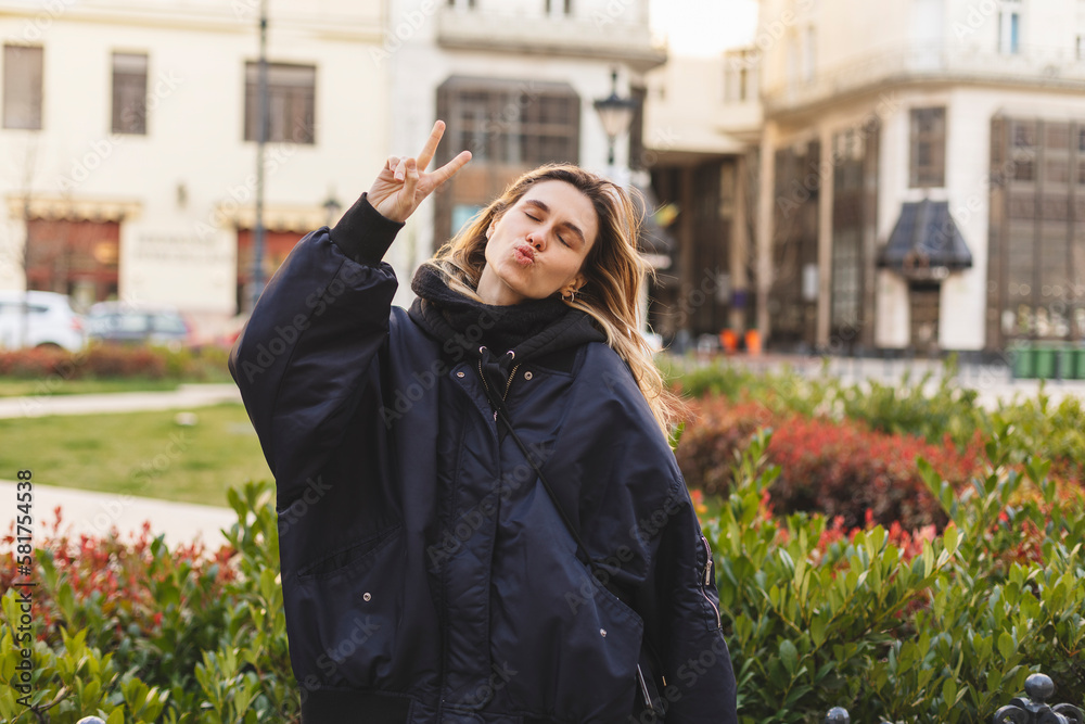 Cool blonde woman in dark blue bomber shows peace v sign, closed eyes, blows kiss and posing outside. Happy woman walk and posing outdoor in the city.