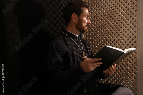 Canvas-taulu Catholic priest in cassock reading Bible in confessional booth