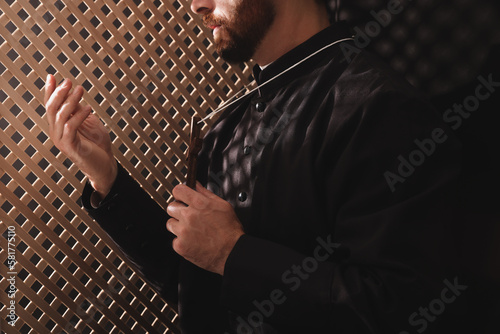 Catholic priest in cassock holding cross and talking to parishioner in confessional, closeup