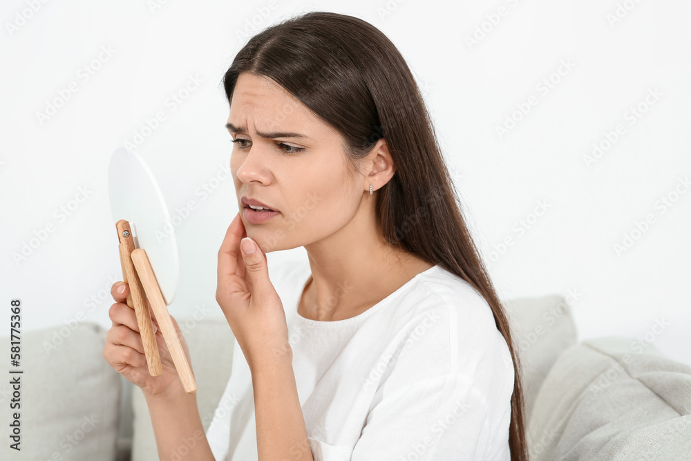 Young woman looking in mirror and touching her face indoors. Hormonal disorders