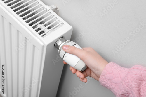 Girl adjusting heating radiator thermostat near white wall indoors, closeup