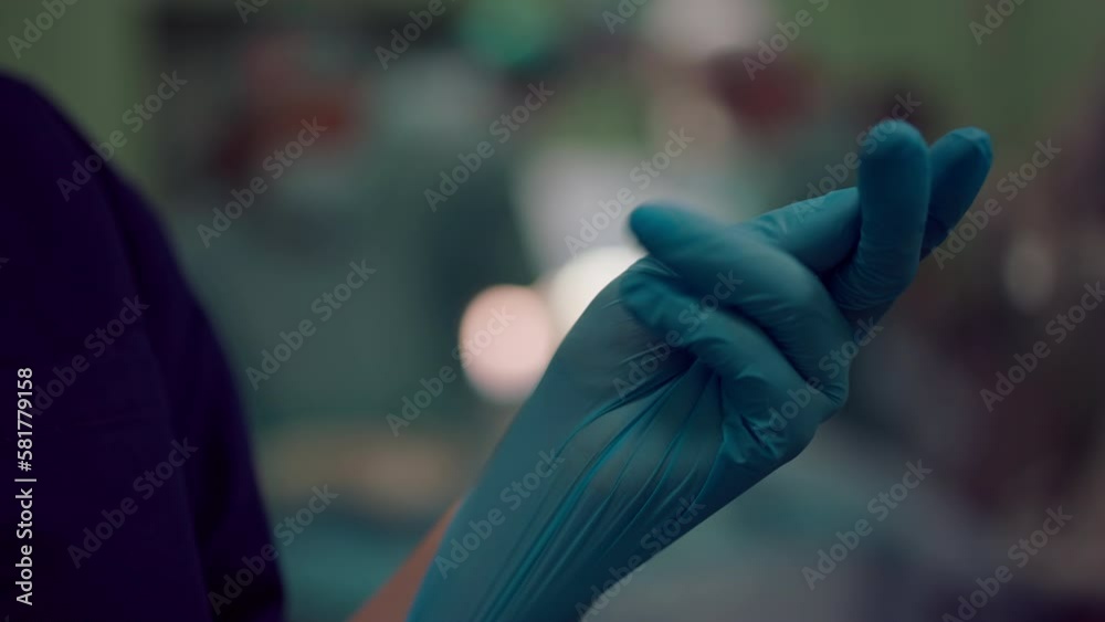 Close-up female hand in rubber medical glove at background of blurred surgery. Unrecognizable woman in sterile surgical gloves in operating room. Slow motion Close-up female hand in rubber medical glove at background of blurred surgery. Unrecognizable woman in sterile surgical gloves in operating room. Slow motion