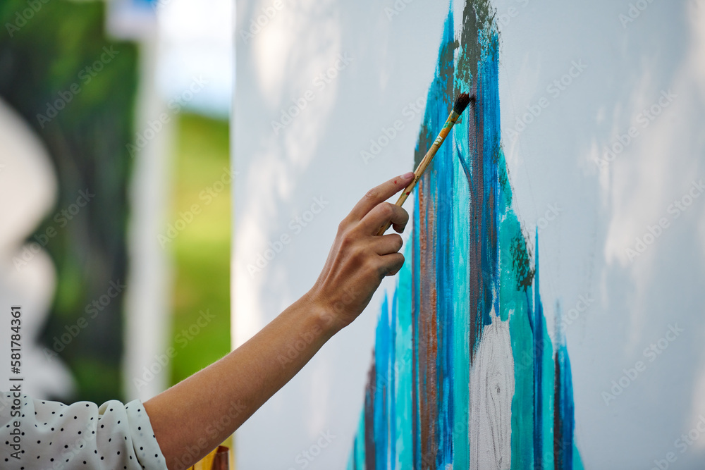Artist hand holds paint brush and draws abstract surreal image on white canvas at outdoor art painting festival, paintings art picture process. Artist paints atmospheric picture - obrazy, fototapety, plakaty 