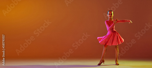 Photos Studio shot of little girl, 11 years old kid in festive stage dress dancing ballroom dance over brown background in neon light
