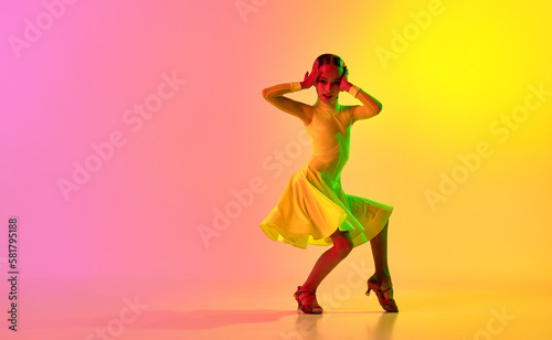 Emotional little girl in yellow stage dress dancing classical ballroom dance over gradient pink-yellow background in neon light filter.