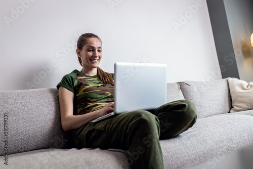 Female soldier making video call using laptop at home while sitting on sofa.