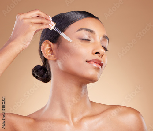 Woman, dropper and retinol on face for skincare beauty or cosmetics against a studio background. Beautiful female with pipette applying oil drop to skin for hydration, moisturizer or facial treatment photo